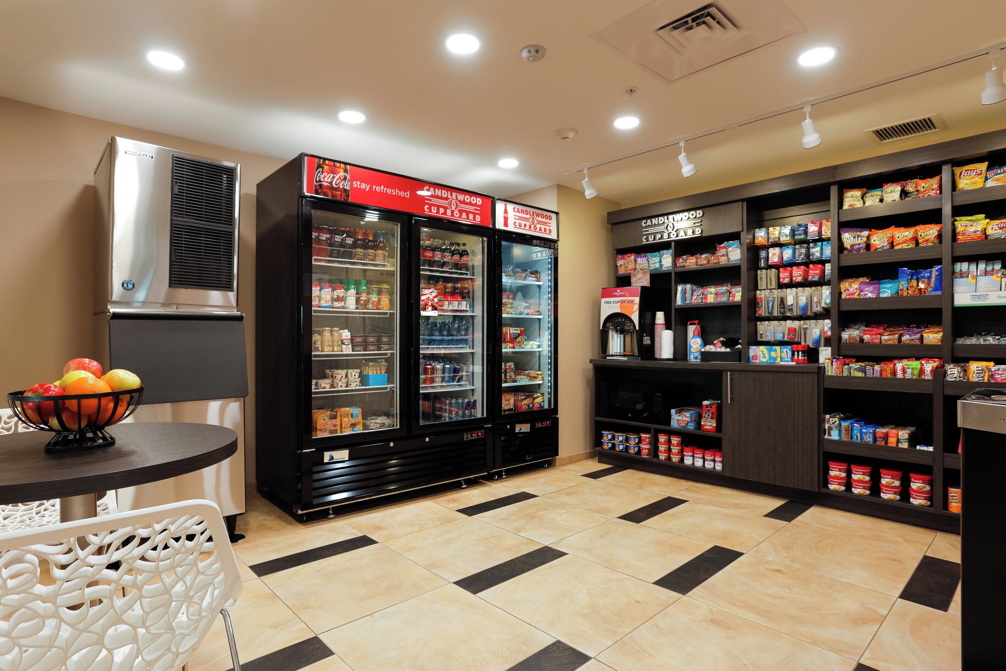 Una kitchenette con refrigerador, dispensador de bebidas y estantes llenos de aperitivos y condimentos.
