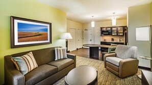 A modern hotel room with a living area, a dining table, a kitchen, and a large window.