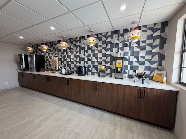 A modern kitchen area with a long counter, coffee machines, a waffle maker, and a wall with a geometric pattern.