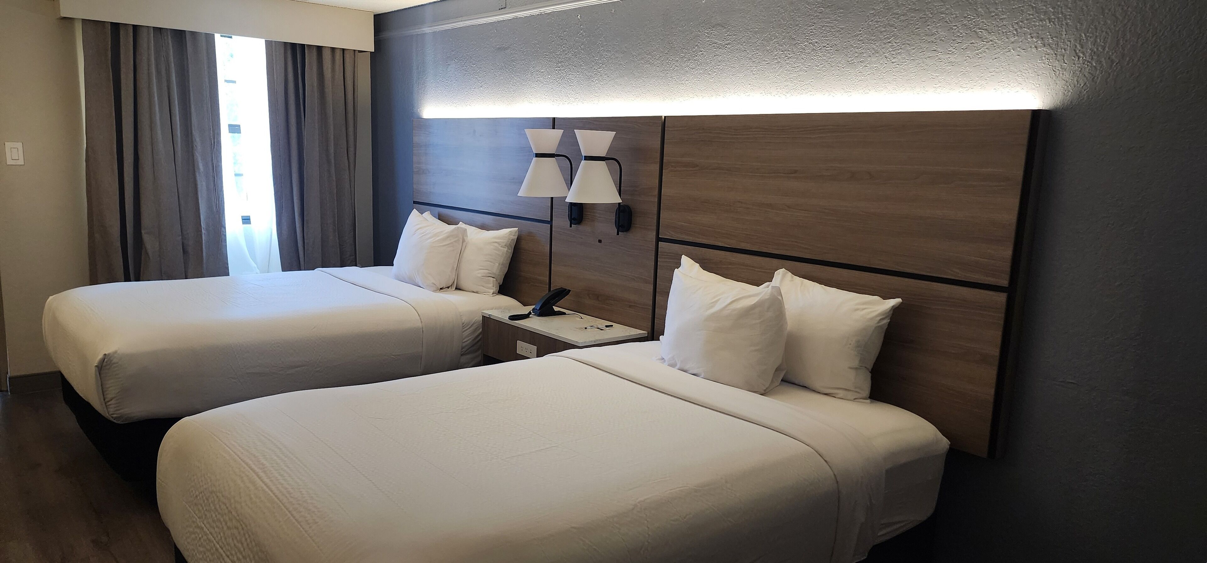 Quarto de hotel com duas camas, uma mesinha de cabeceira, uma luminária e vista para o exterior pela janela.