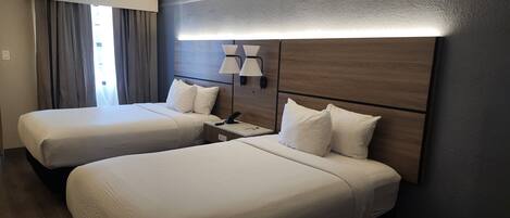 Quarto de hotel com duas camas, uma mesinha de cabeceira, uma luminária e vista para o exterior pela janela.