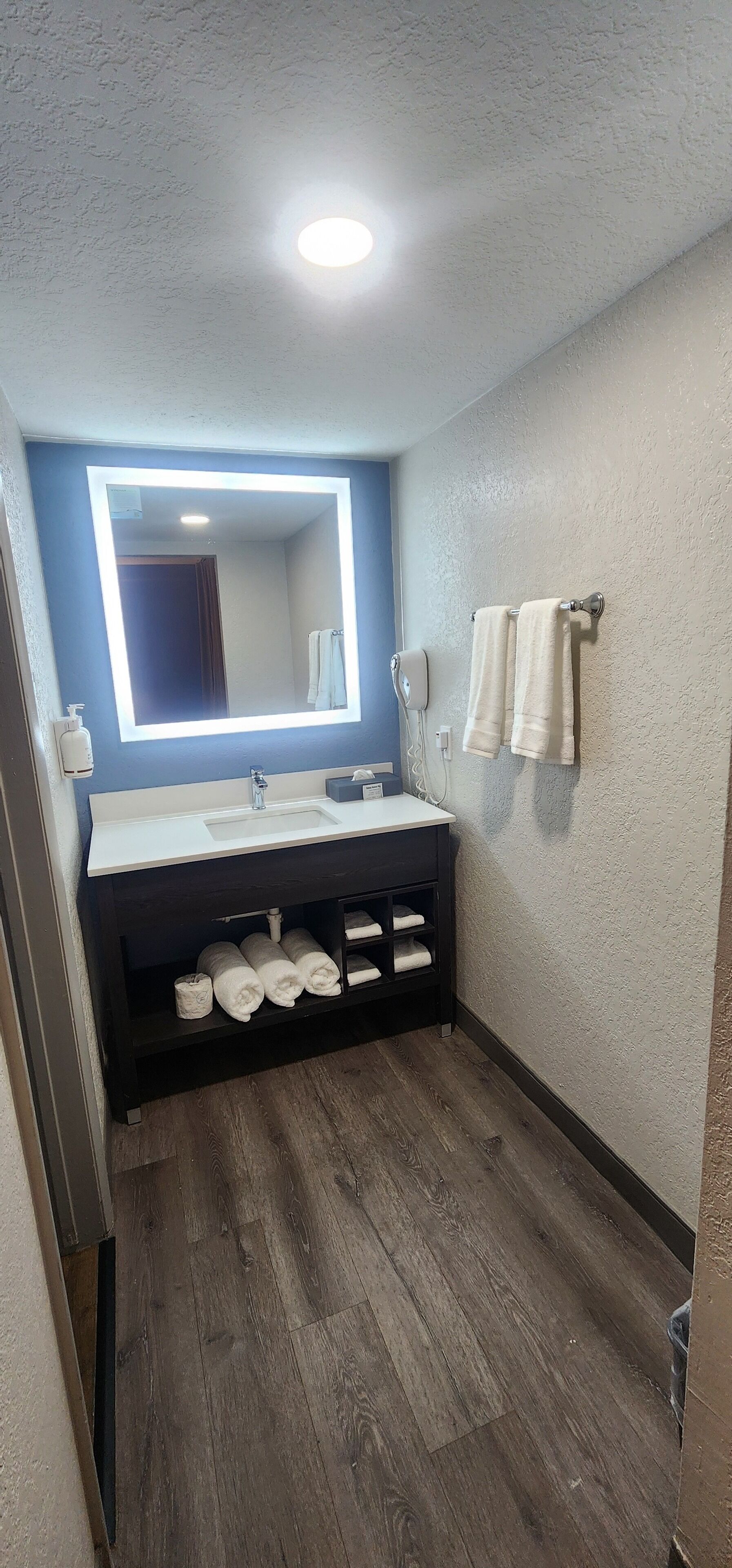 A bathroom with a white vanity, a large mirror with underglow lighting, two towels on a wall-mounted rack, and a wooden floor.