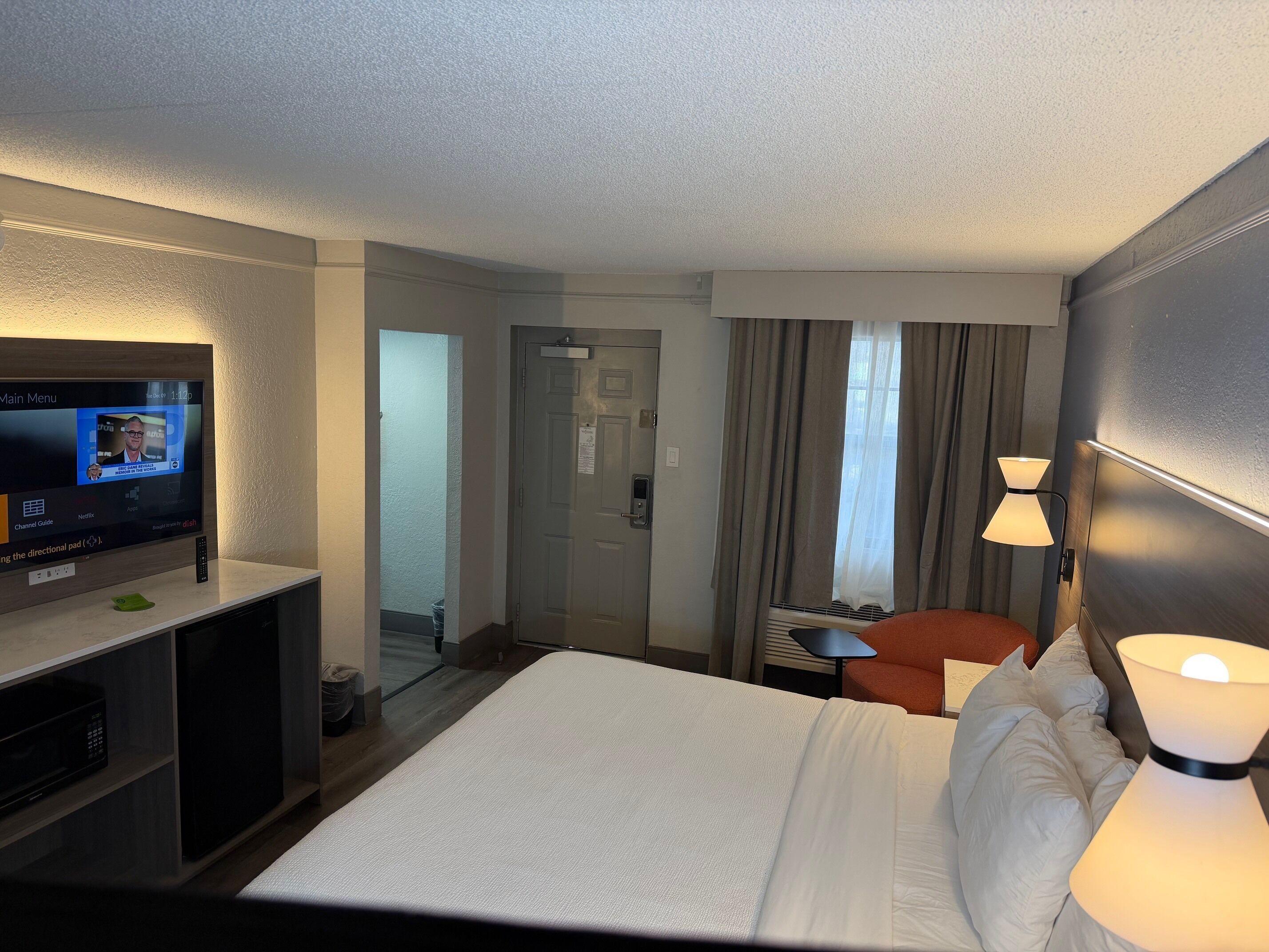 A hotel room with a bed, a TV on a stand, a desk with a chair, and a lamp.