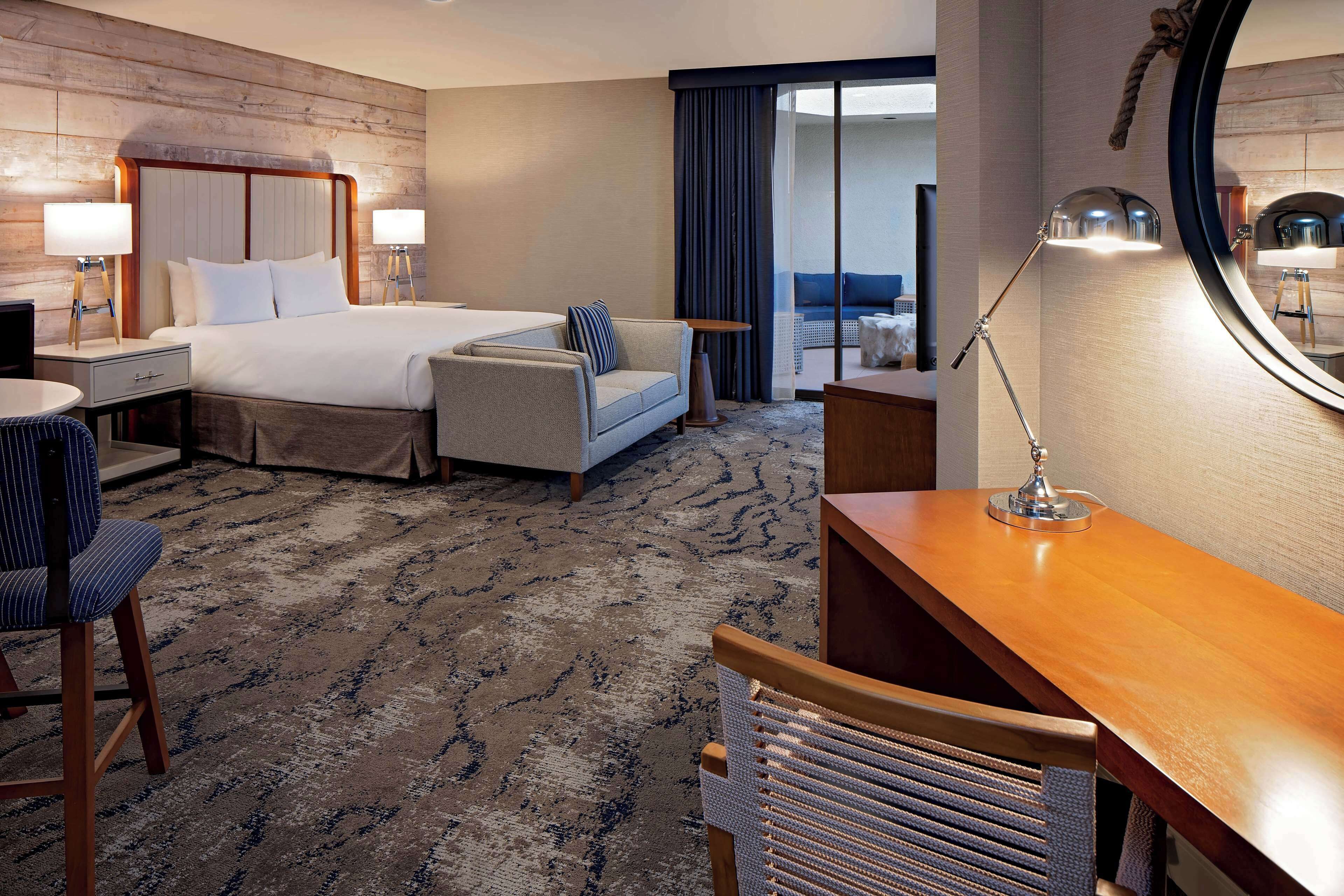 A hotel room with a large bed, a sofa, a desk, and a chair. There is a mirror on the wall and a window with curtains.