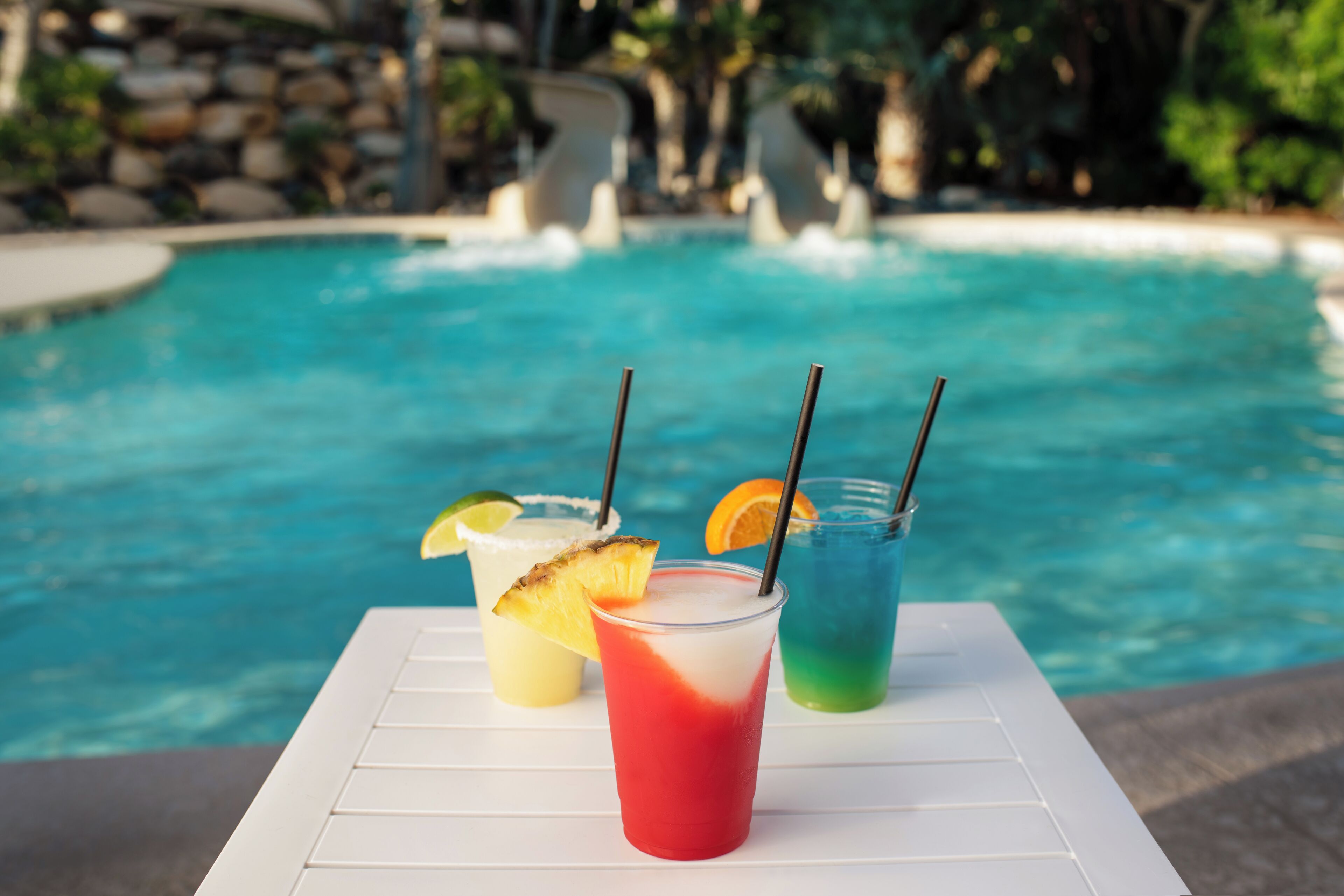 A poolside table with three drinks, each with a straw and garnished with a slice of fruit.