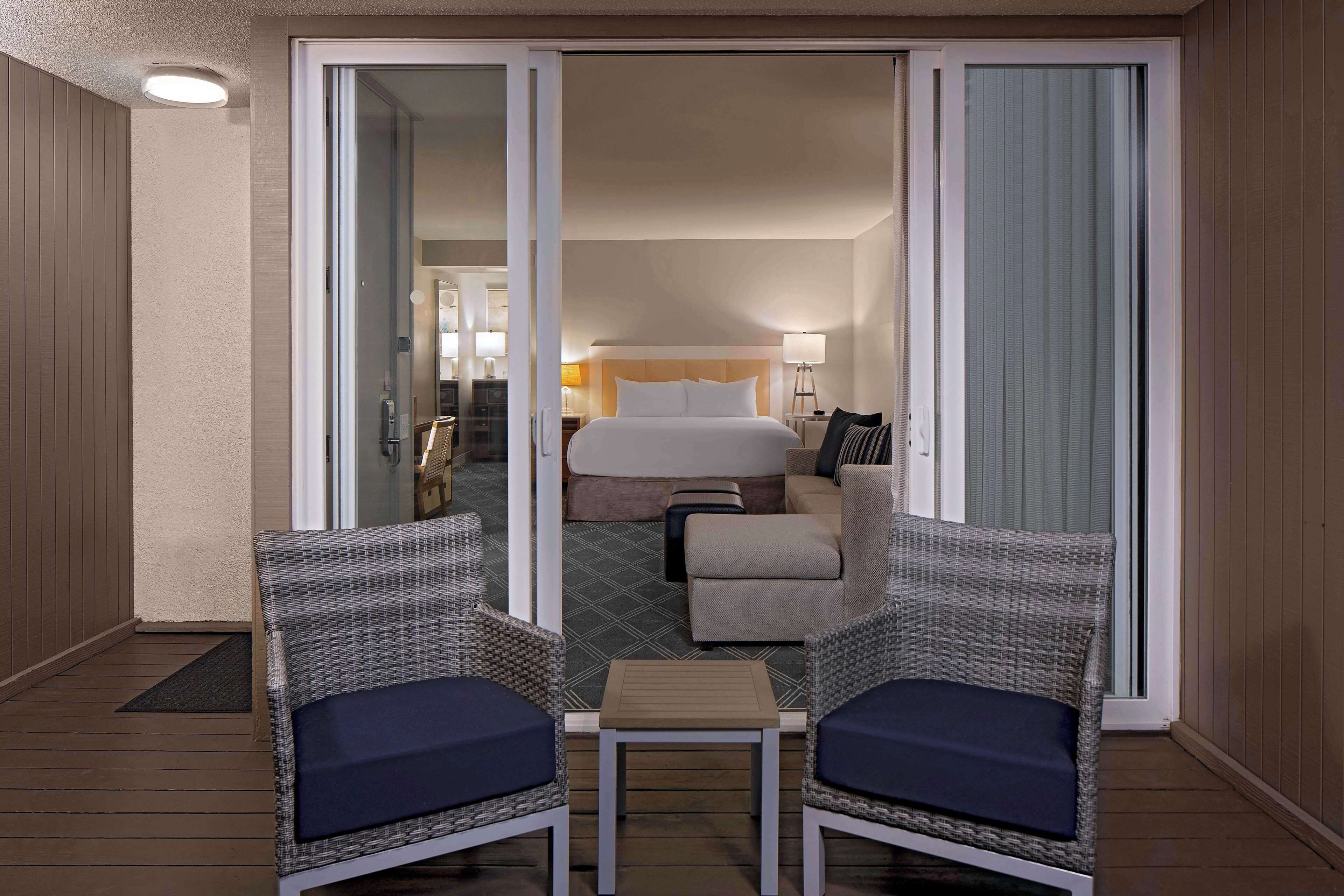 A hotel room with a bed, a small table, two chairs, and a view through a sliding door.