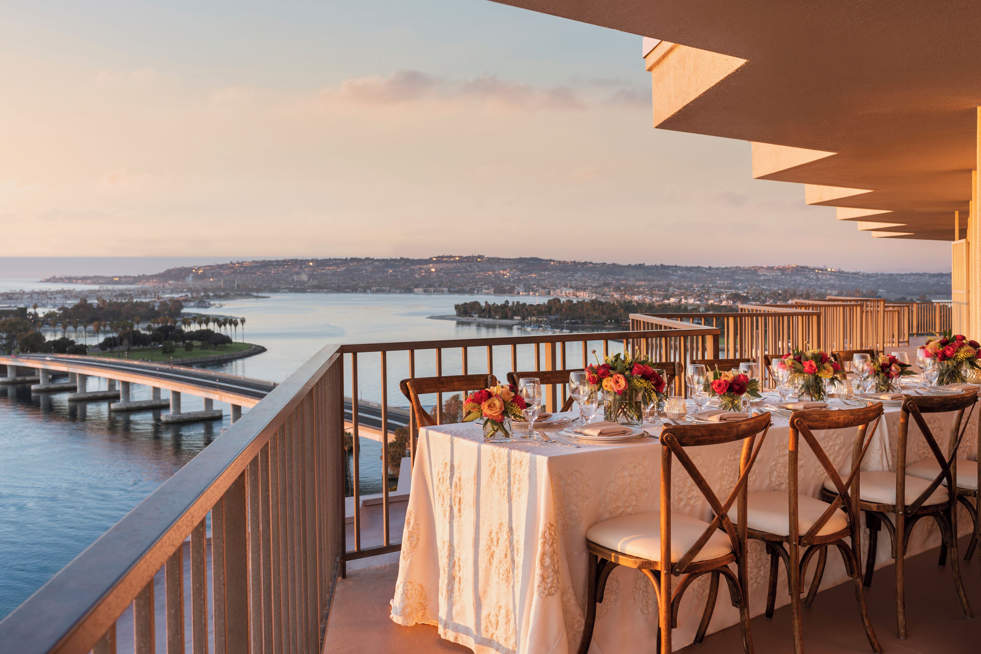 A dining table set for six with chairs, overlooking a body of water and a bridge.