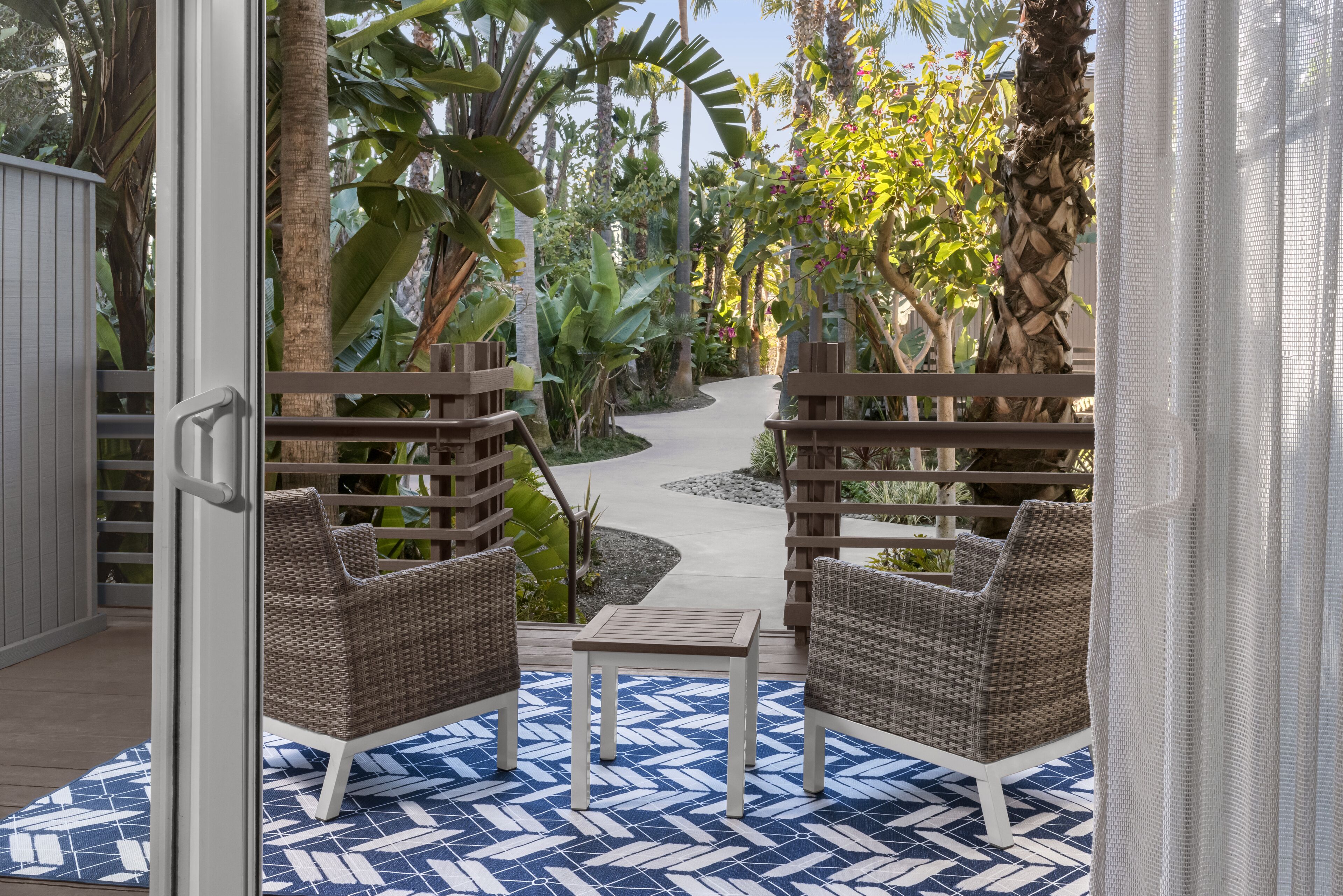 Eine Terrasse mit Korbstühlen und einem kleinen Tisch, umgeben von üppiger tropischer Bepflanzung und einem Weg.