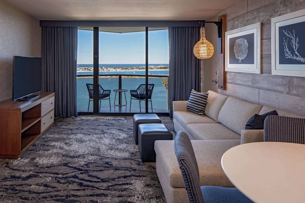Una camera d'hotel moderna con una grande finestra che offre vista sul mare, un divano, un tavolo da pranzo e una TV su un mobile in legno.