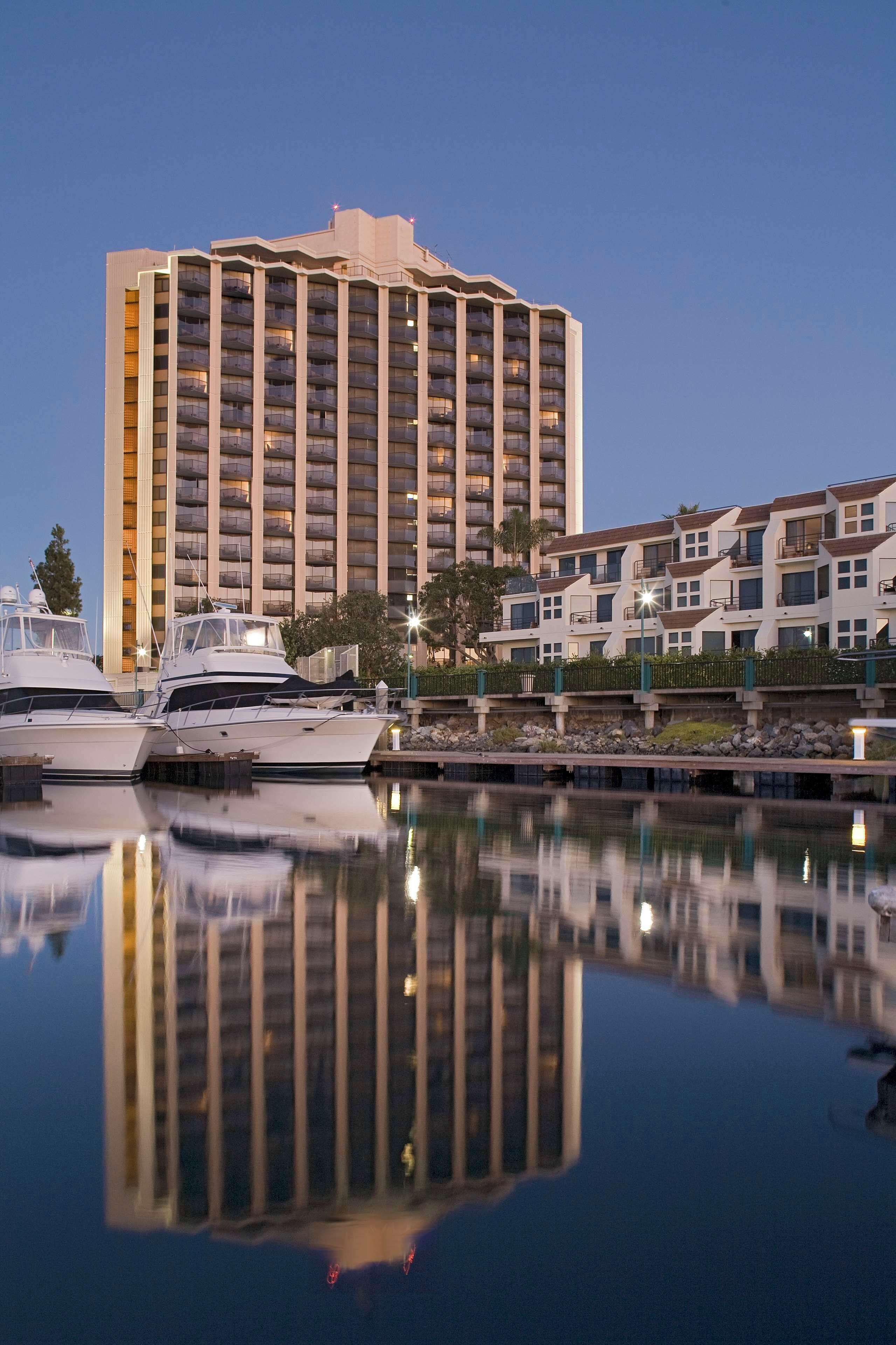 A high-rise hotel with multiple balconies, a marina with boats, and a calm body of water reflecting the buildings.