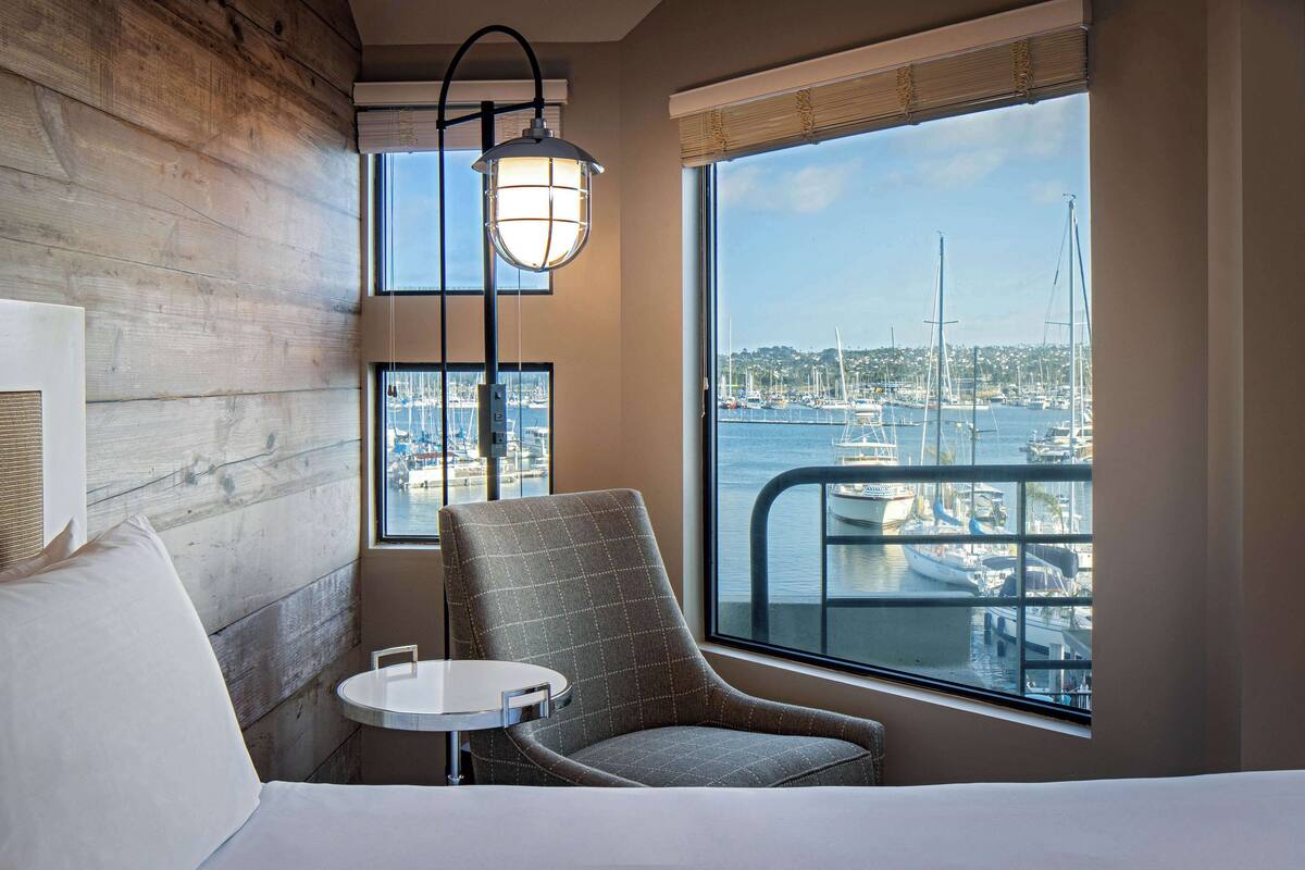 Camera d'hotel con una grande finestra vista sul porto turistico, una poltrona comoda, un tavolino e un letto.