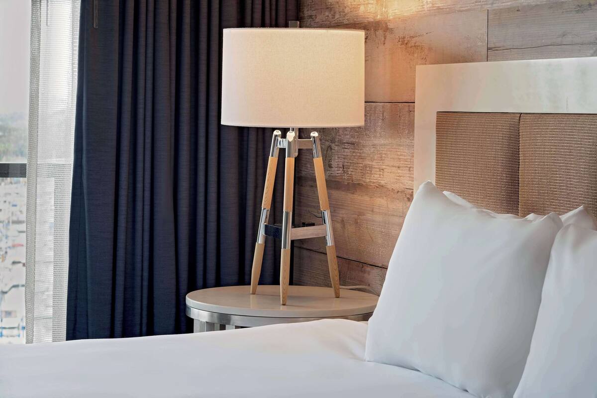Una camera d'albergo con un letto, un tavolino rotondo, una lampada su un supporto a treppiede e vista sulla città dalla finestra.
