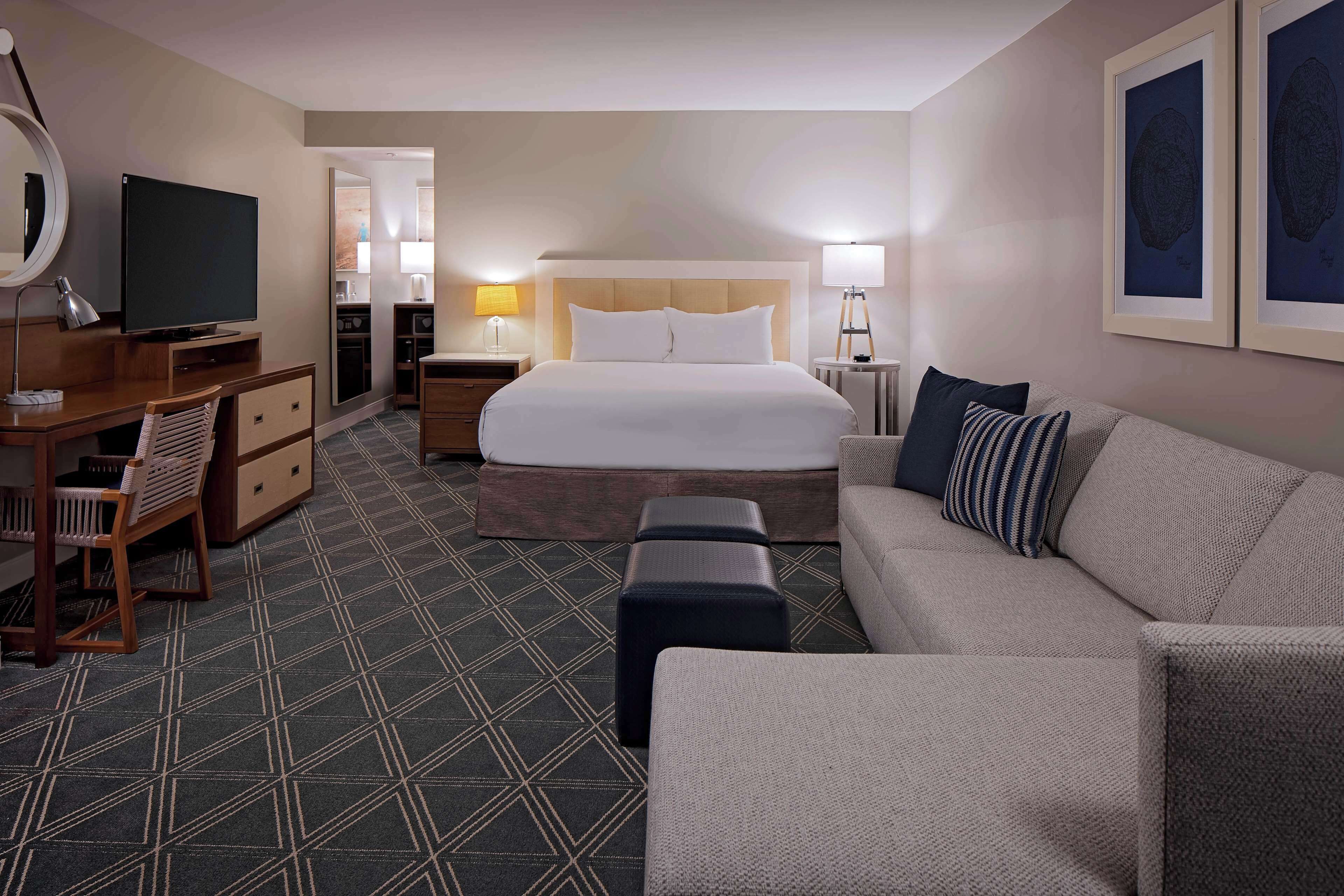 A hotel room with a large bed, a sitting area with a sofa and armchair, a desk with a computer, a TV, and a mirror.