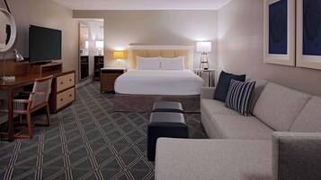 A hotel room with a large bed, a sitting area with a sofa and armchair, a desk with a computer, a TV, and a mirror.