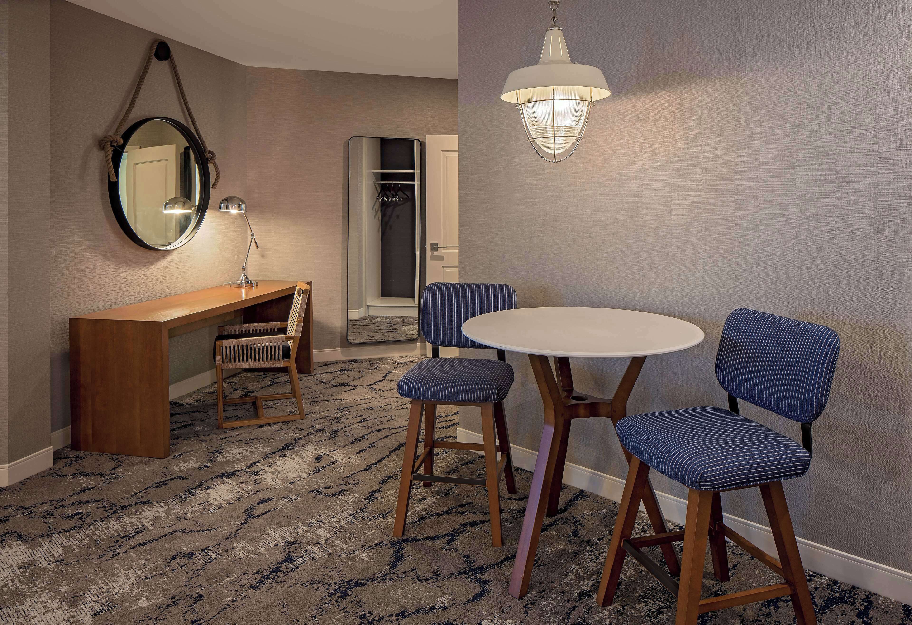 Quarto de hotel com uma mesa redonda, duas cadeiras estofadas, uma escrivaninha de madeira, um espelho e um lustre.
