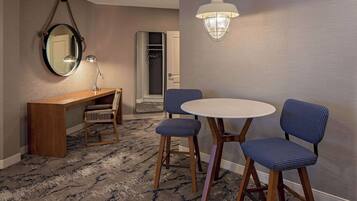 Quarto de hotel com uma mesa redonda, duas cadeiras estofadas, uma escrivaninha de madeira, um espelho e um lustre.