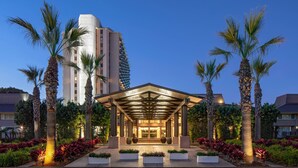 Una entrada de hotel con un corredor cubierto, palmeras y un edificio moderno al fondo.