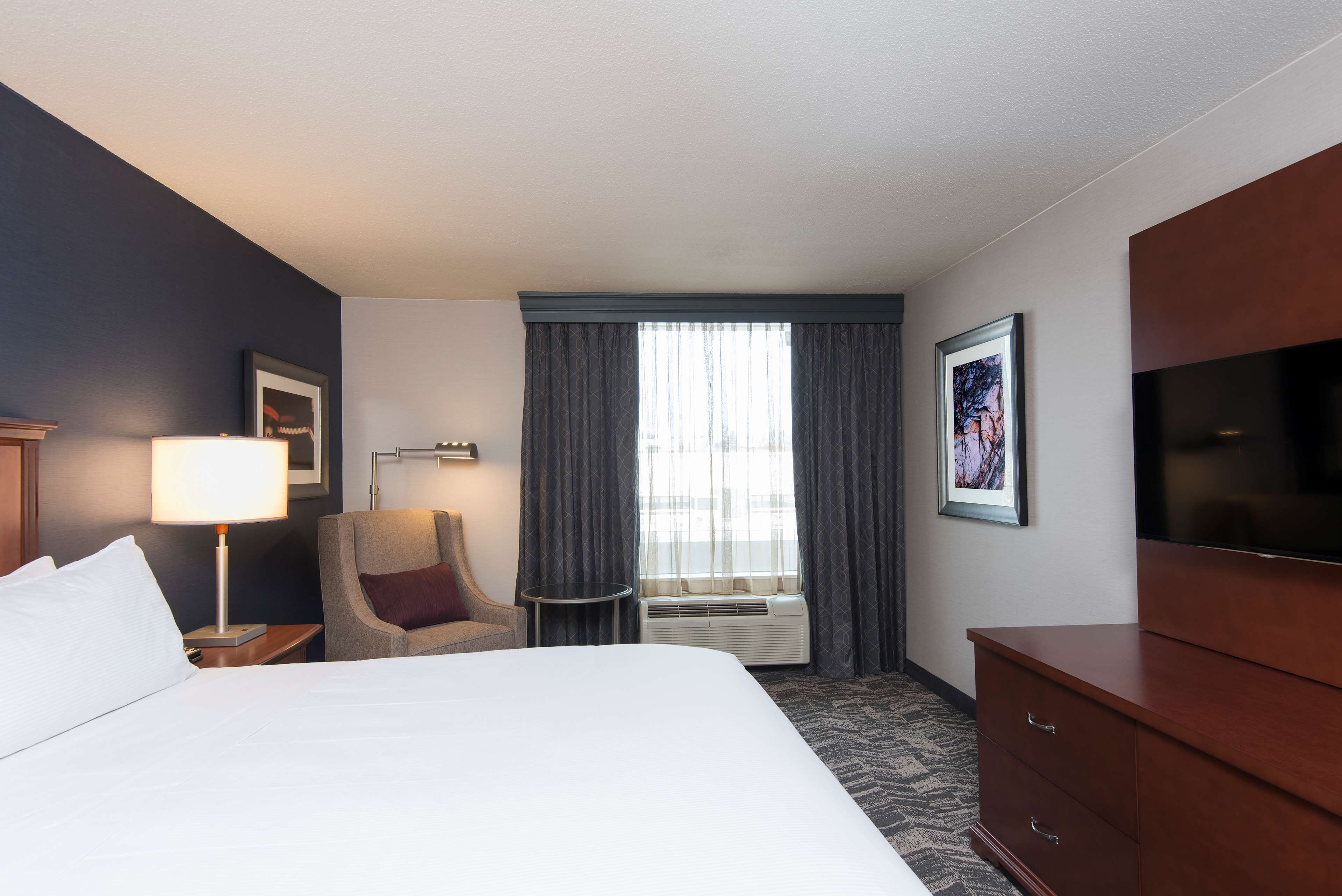 Habitación de hotel con una cama grande, una silla, un escritorio con lámpara, un televisor y una ventana con cortinas.