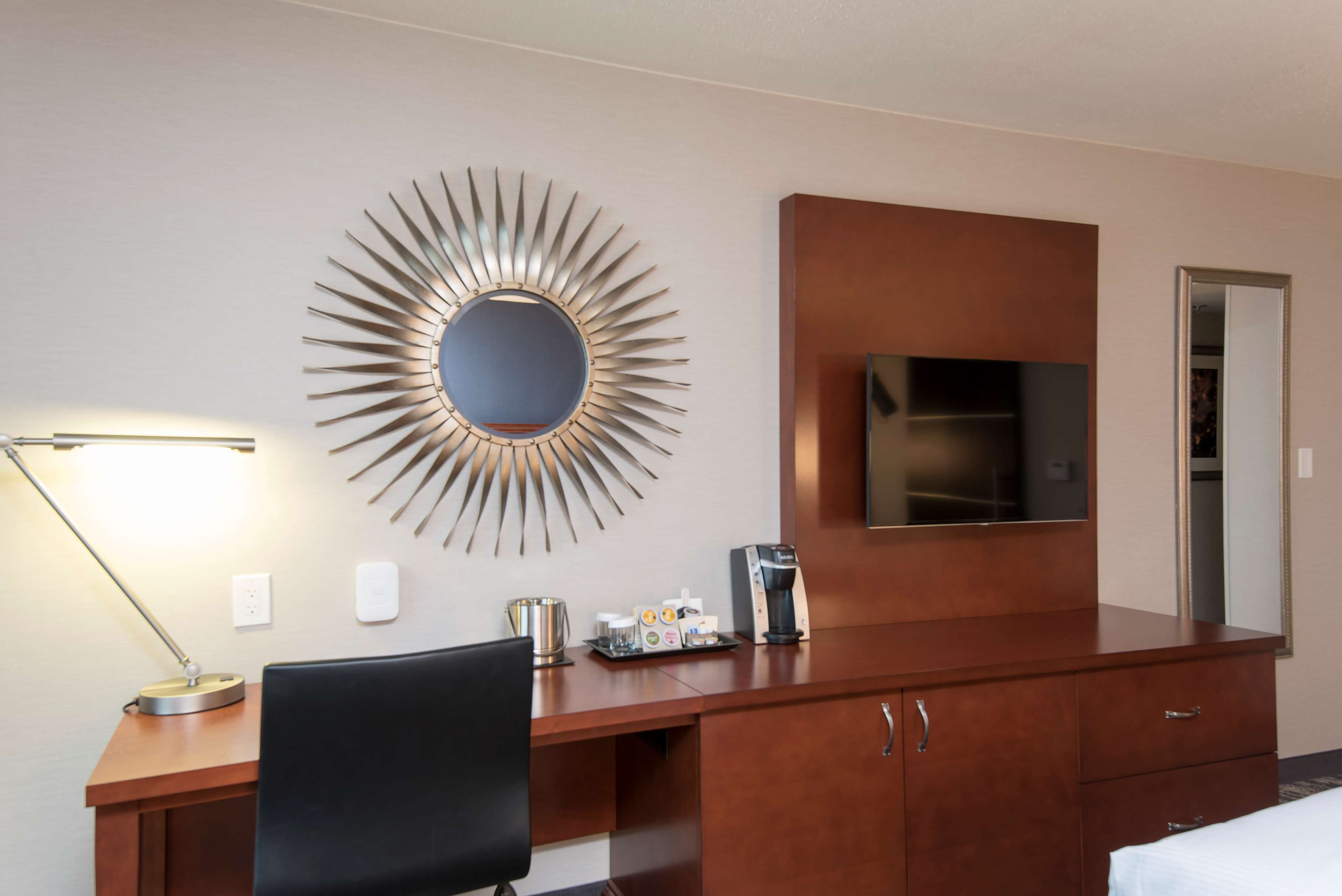 Una camera d'hotel con una scrivania, una sedia da ufficio nera, una macchina del caffè, una TV fissata al muro e uno specchio con motivo a raggiera.