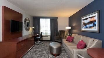 A hotel room with a flat-screen TV, a sofa, a round ottoman, a desk with a chair, and a window with curtains.