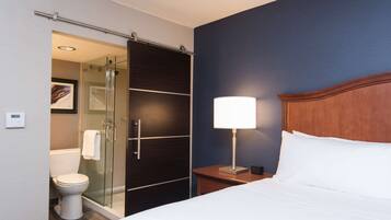 Habitación de hotel con cama, mesita de noche con lámpara, cabecera de madera, cabina de ducha de vidrio, inodoro y un cuadro enmarcado en la pared.