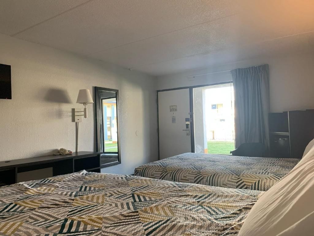 A hotel room with two beds, a desk, a lamp, and a view of the outside.