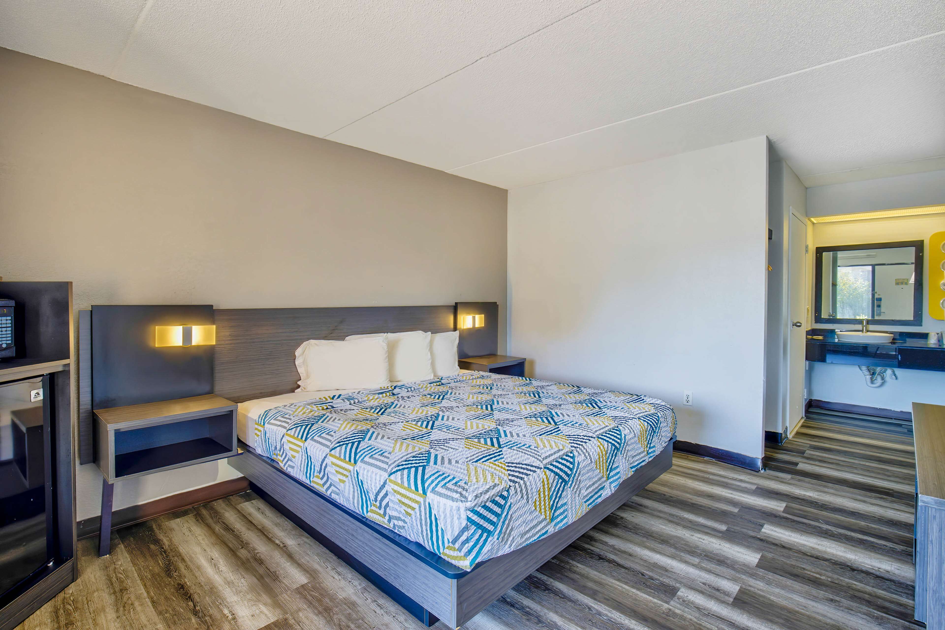 A modern hotel room with a large bed, bedside tables, a built-in TV, and a bathroom with a mirror and sink.
