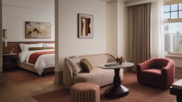 A hotel room with a bed, a sofa, a red armchair, a small table, and a window with a view of buildings.