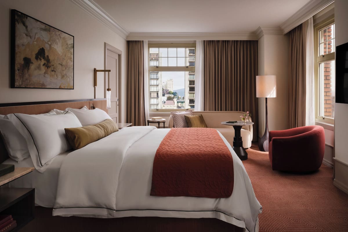 Habitación de hotel con una cama grande, un sofá, una mesita y una silla roja. Hay una ventana con cortinas, un cuadro en la pared y una lámpara.