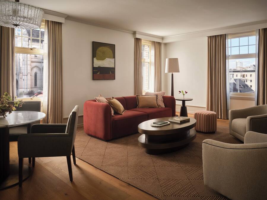 A modern living room with a red sofa, a round coffee table, a lamp, and a view of buildings through large windows.
