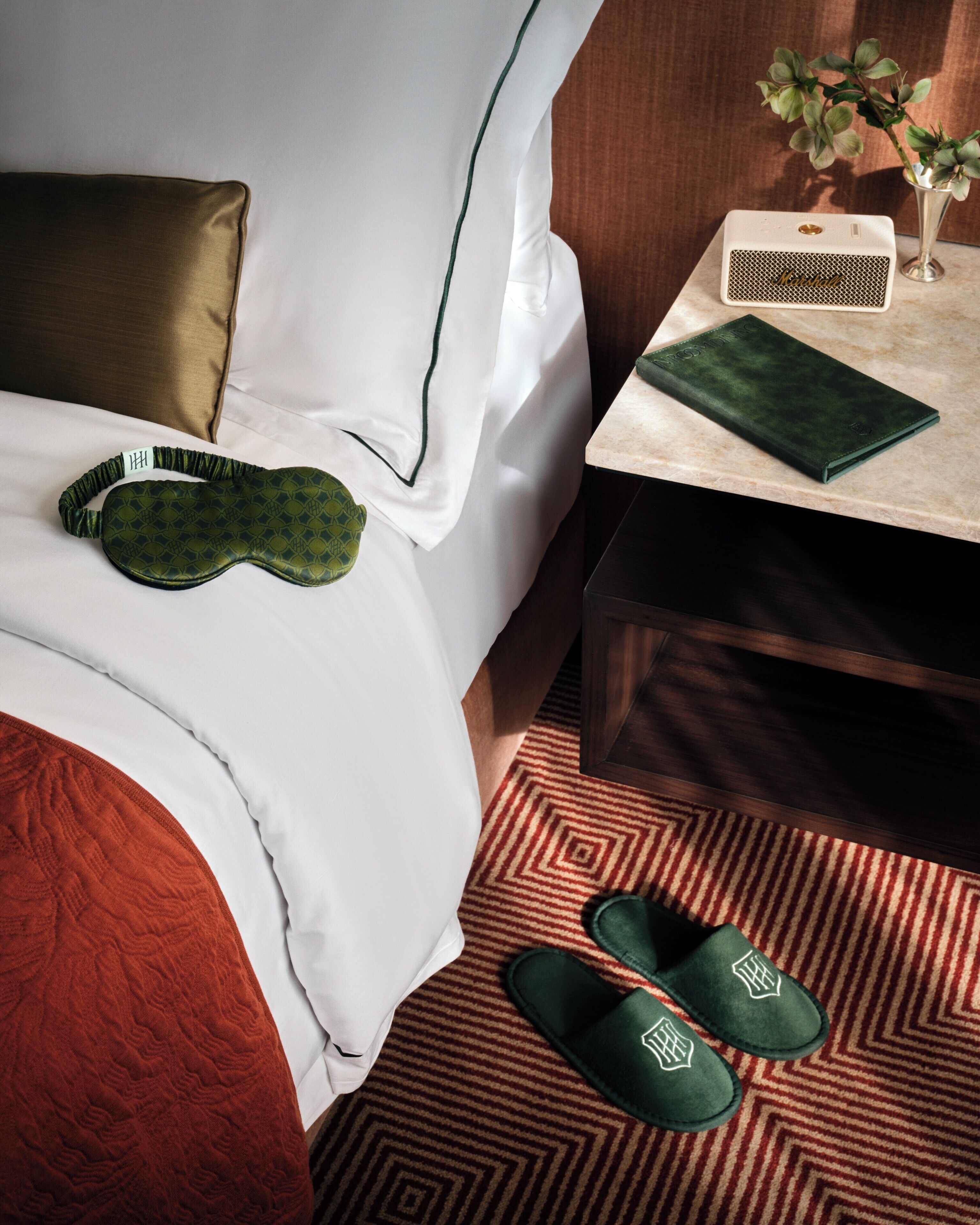 Un lit bien fait, agrémenté d’un masque de sommeil vert, d’une paire de chaussons verts, d’une petite table avec un haut-parleur et d’un vase contenant des feuilles vertes.