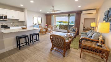 A modern kitchen with a microwave, stove, and bar stools. There is a dining area with wicker chairs and a sofa. The room has a large window with a view of the ocean.