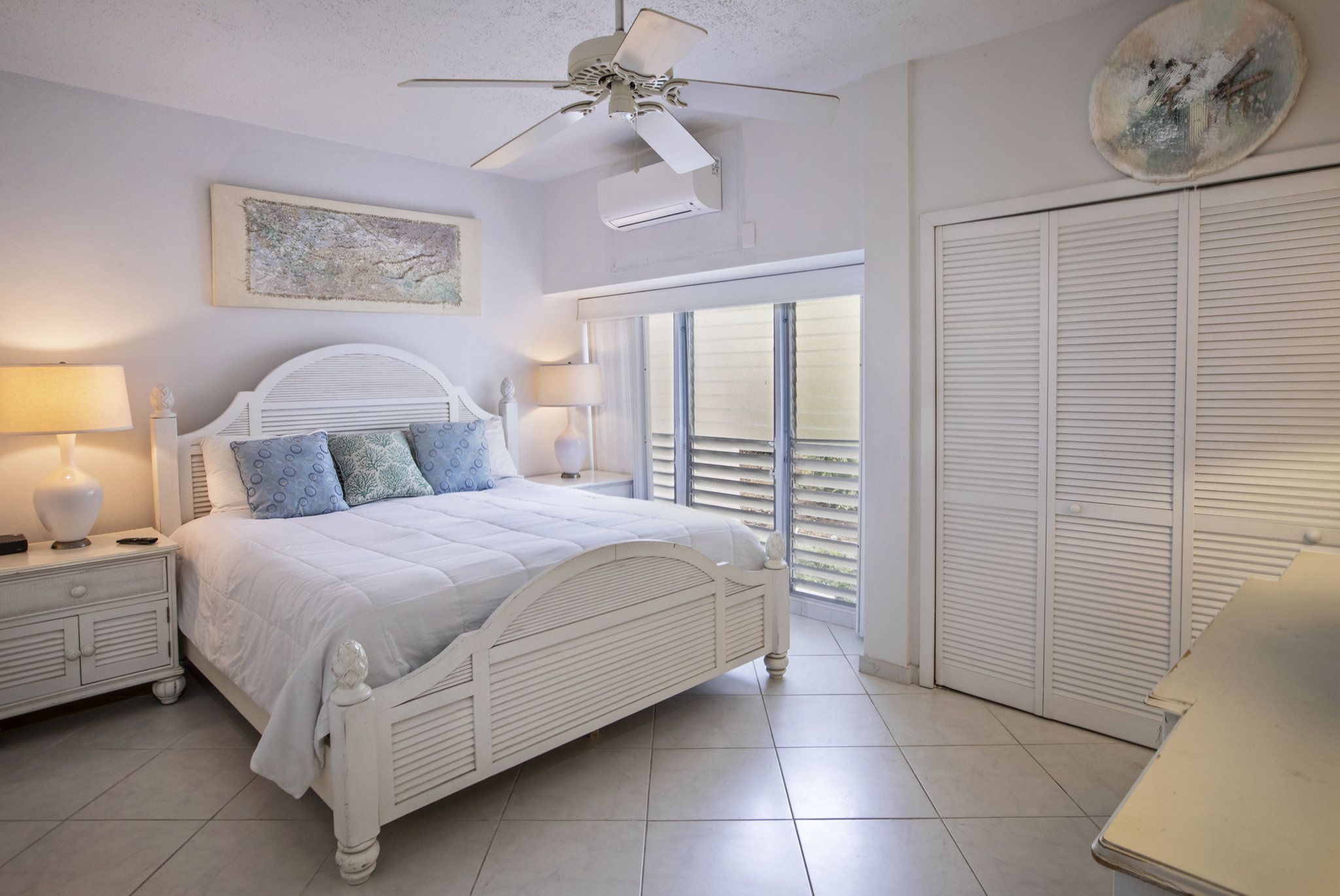 A bedroom with a white bed, two bedside tables with lamps, a ceiling fan, a window with blinds, and a closet.