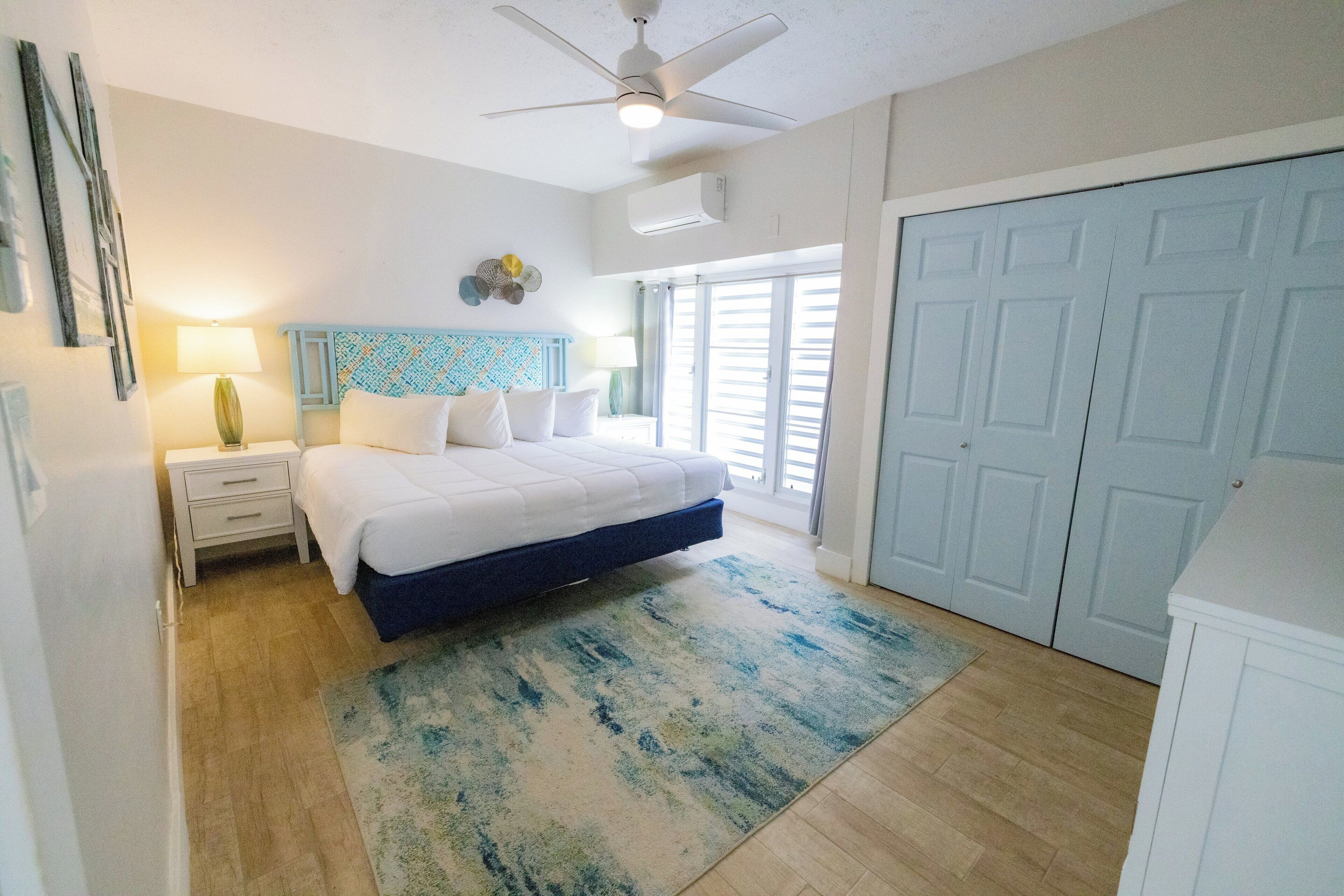A bedroom with a bed, nightstands, a ceiling fan, a window with blinds, and a door.
