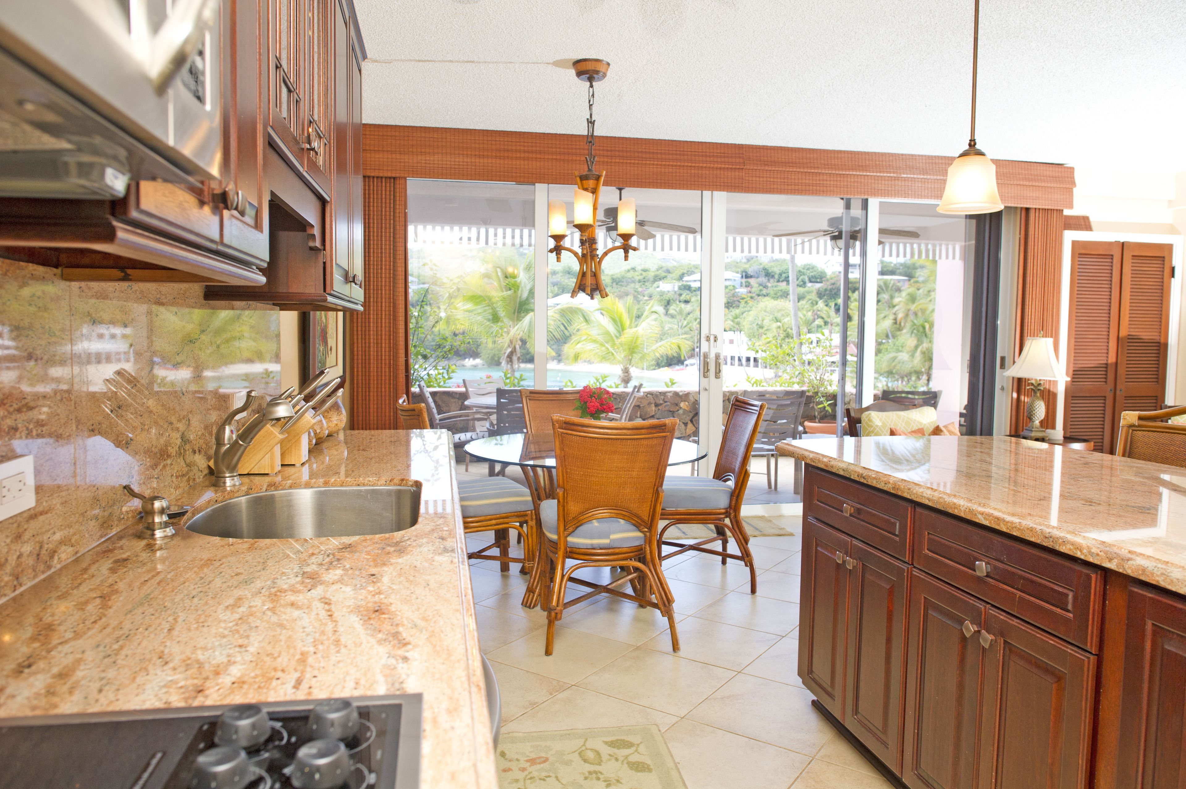 Una cocina con encimeras de granito, armarios de madera, un comedor con sillas de mimbre y vistas a un paisaje tropical a través de amplias ventanas.