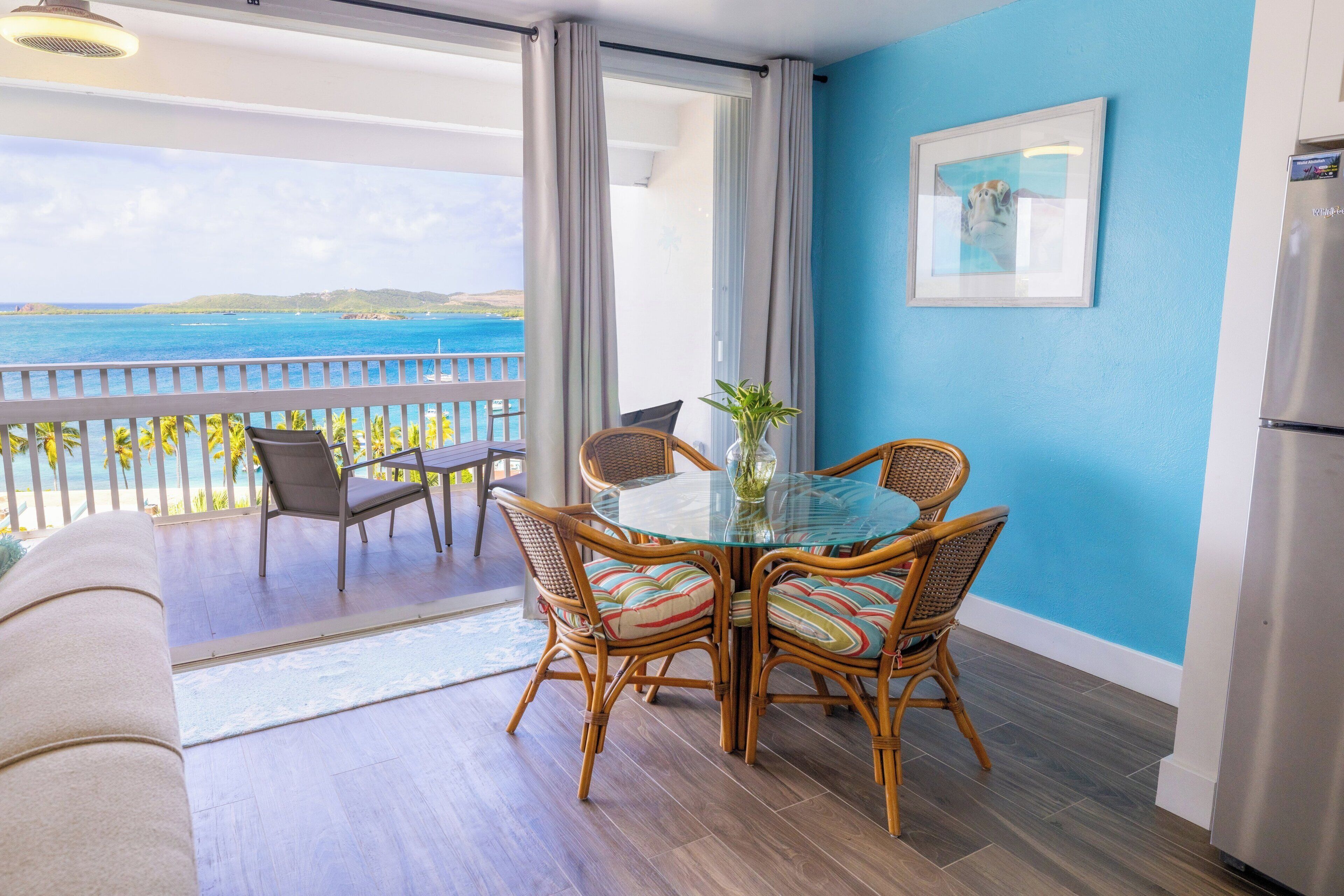 A balcony with a view of the sea, a dining table with wicker chairs, and a refrigerator.