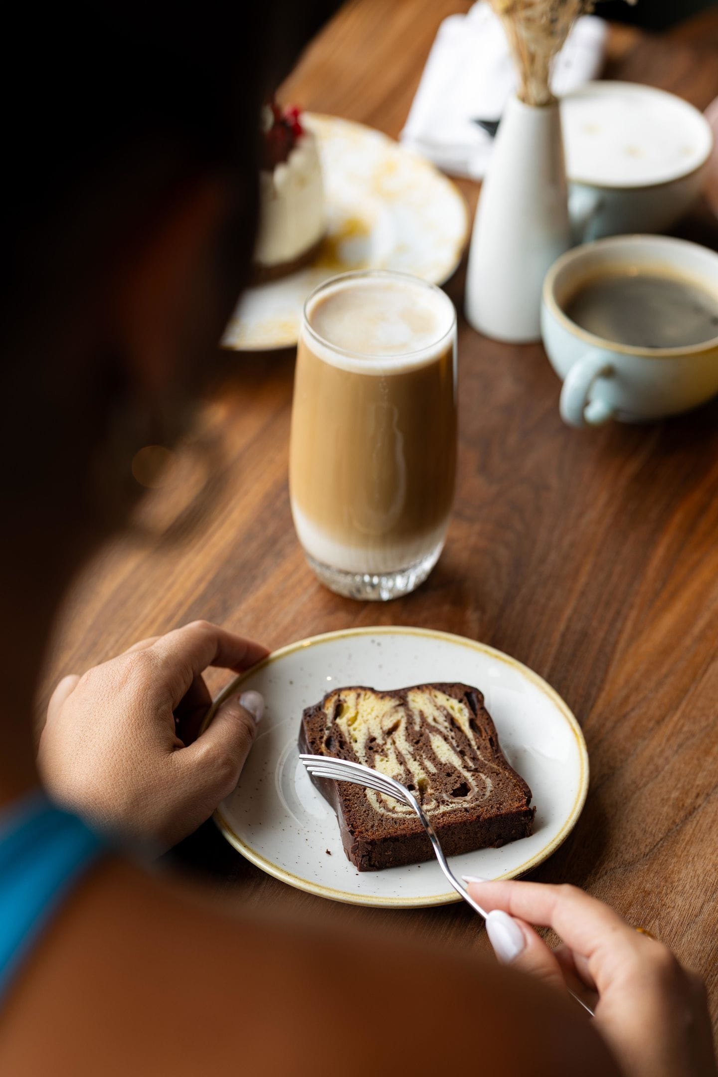 Una persona comiendo una porción de pastel con un tenedor sobre un plato, una taza de café y una taza en una mesa de madera.