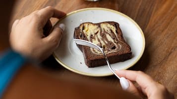 Una persona comiendo una porción de pastel con un tenedor sobre un plato, una taza de café y una taza en una mesa de madera.