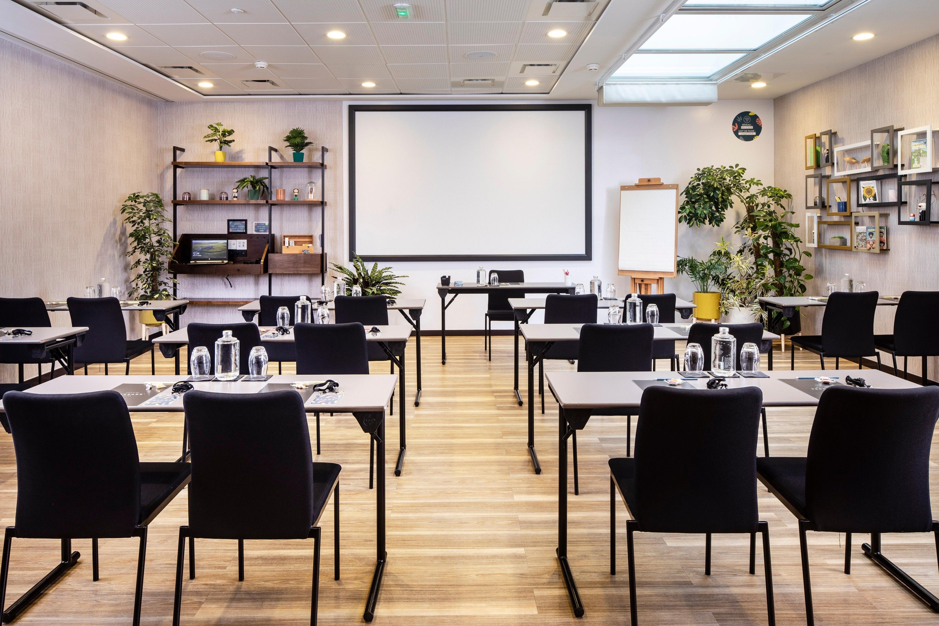 Une salle de conférence dotée d’un grand écran, de plusieurs tables, de chaises et d’étagères ornées de plantes et d’objets décoratifs.