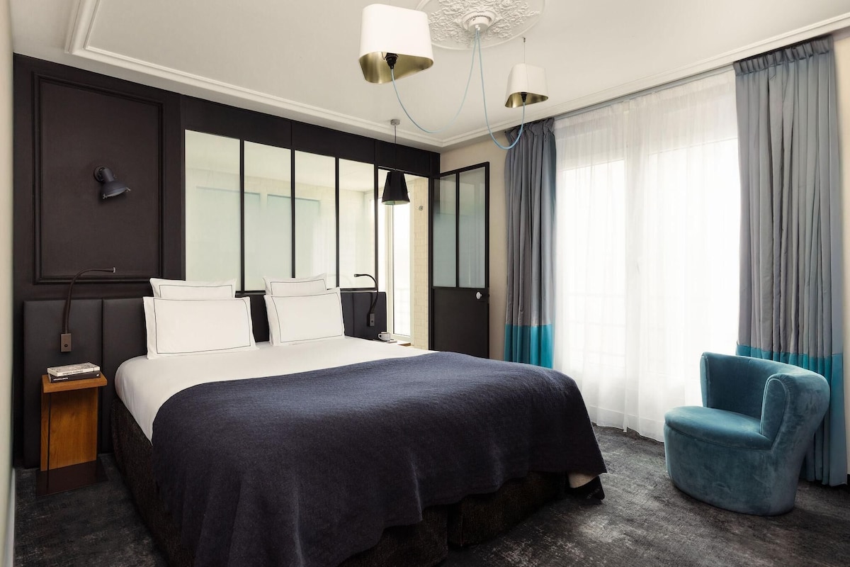 A modern hotel room with a large bed, a side table, a chair, and a large window with curtains.