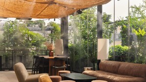 Un espacio exterior moderno con zona de estar, sofá, sillas y una mesa, rodeado de paredes de vidrio y vegetación.