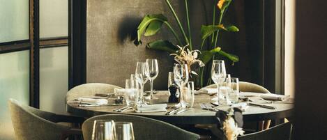 Una mesa de comedor dispuesta con vasos y cubiertos, rodeada de sillas, con una planta grande al fondo.