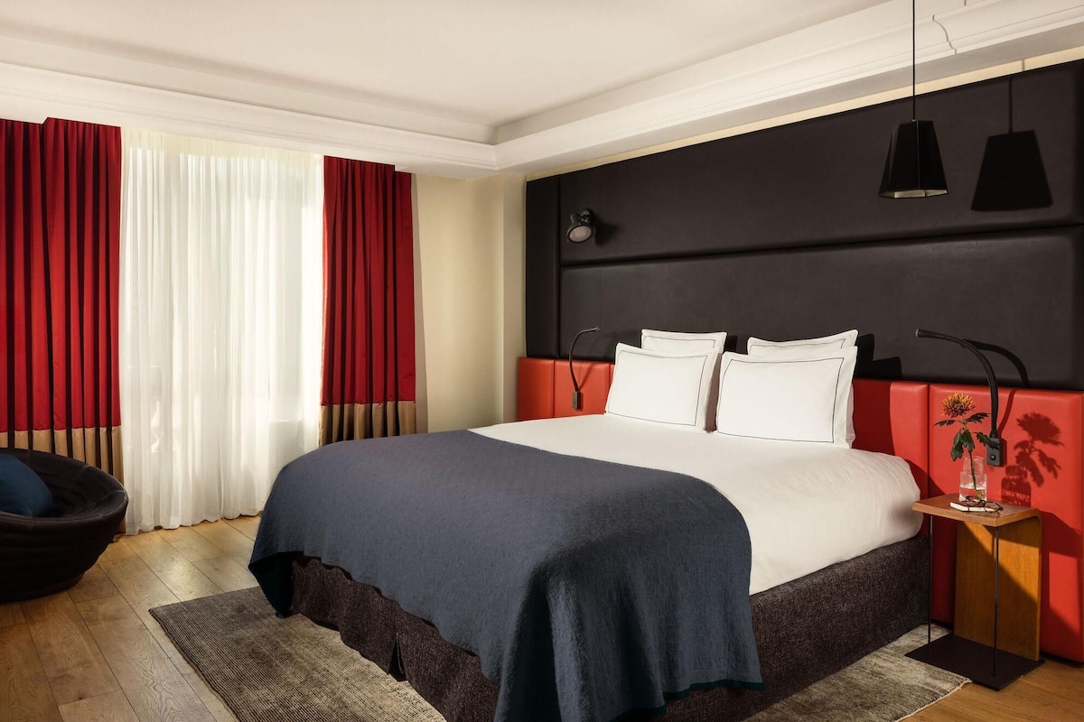 A modern hotel room with a large bed, red curtains, a black headboard, and a small bedside table with a vase.