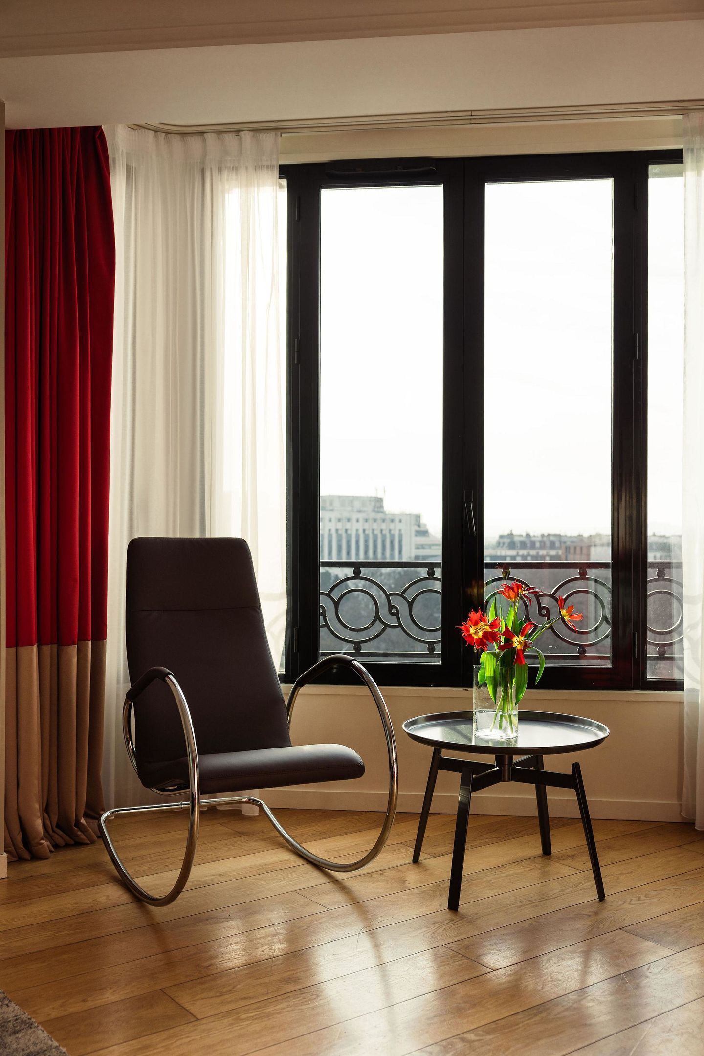 A room with a black chair, a small round table with a vase of flowers, and a view through large windows.