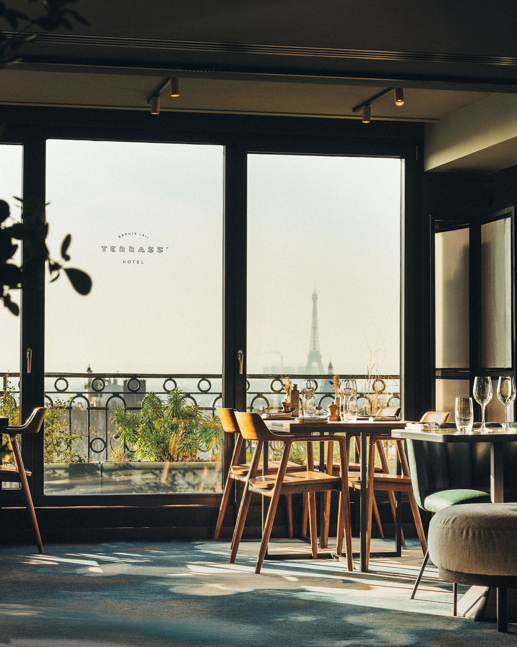 A dining area with a view of the Eiffel Tower, featuring a table set for two with chairs, a balcony railing, and potted plants.