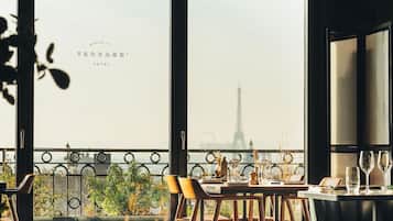 A dining area with a view of the Eiffel Tower, featuring a table set for two with chairs, a balcony railing, and potted plants.