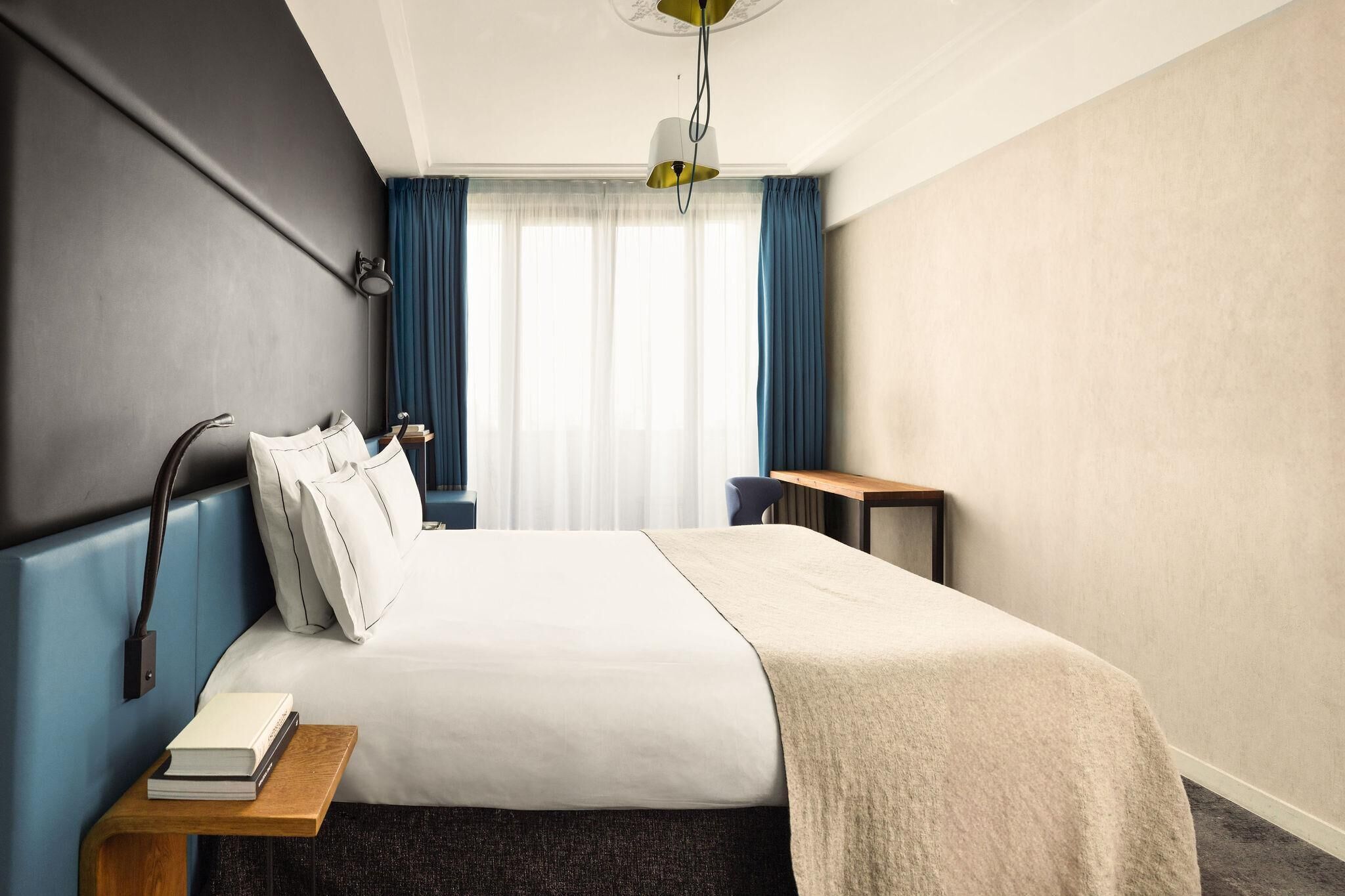 Habitación de hotel con cama, mesita de noche, lámpara y vistas al exterior a través de una ventana con cortinas.