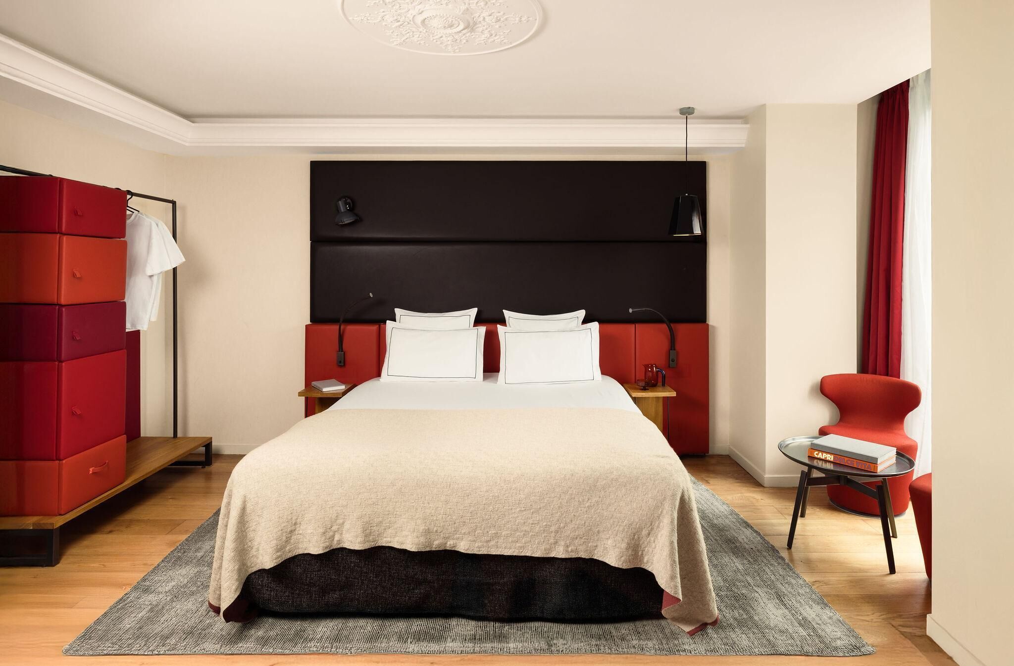 Ett modernt hotellrum med en stor säng, en röd garderob, ett litet bord med en bok och en röd fåtölj.