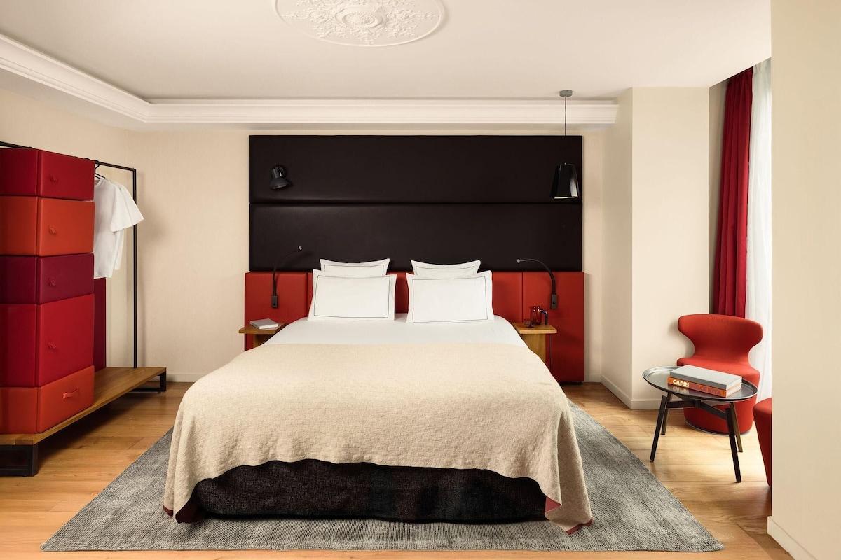 A modern hotel room with a large bed, a red wardrobe, a small table with a book, and a red armchair.