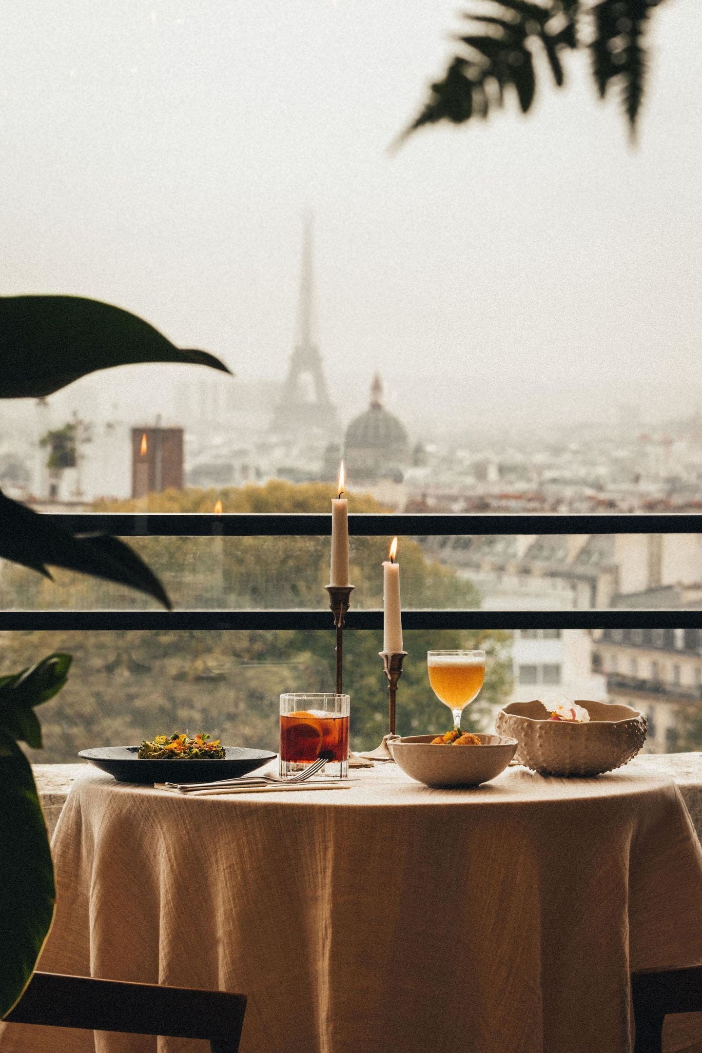 A table with a candle, a glass of amber liquid, and a bowl of food, set against a backdrop of a cityscape with a prominent tower.